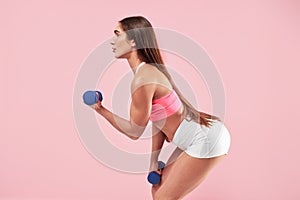 Brunette woman with muscular body in sports clothing training and doing workout with dumm bells on pink background.