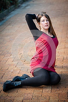 Brunette woman in a red dress with black stockings sitting on a brick walkway