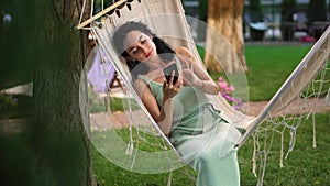 Brunette woman relaxing in a hammock outdoors, surfing internet in her smartphone, clicks on the screen