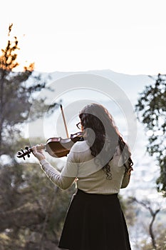 Brunette woman seen from back playing violin outside in the mountain. Vertical