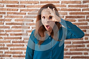 Brunette woman standing over bricks wall doing ok gesture shocked with surprised face, eye looking through fingers