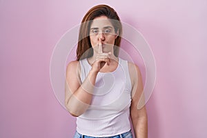 Brunette woman standing over pink background asking to be quiet with finger on lips