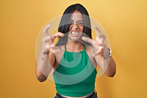 Brunette woman standing over yellow background shouting frustrated with rage, hands trying to strangle, yelling mad