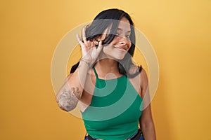 Brunette woman standing over yellow background smiling with hand over ear listening an hearing to rumor or gossip