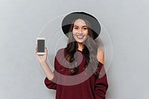 Brunette woman in sweater and hat showing blank smartphone screen