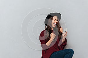 Brunette woman in sweater and hat using smartphone and rejoice