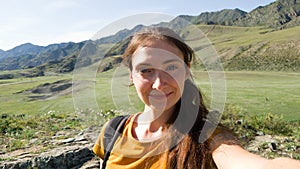 A brunette woman takes a selfie against the background of a field and mountains. Green summer valley. Traveling through a beautifu