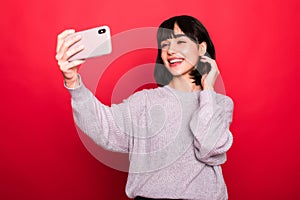 Brunette woman taking selfie with victory sign on her cell phone isolated over red background