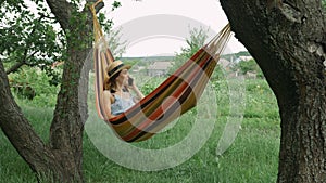 Brunette woman talking by mobile phone in hammock outdoors. Young girl speaking by smartphone while lying in hammock between two t