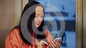 Brunette woman texting message in mobile phone on window background. Adult woman using smartphone for mobile sms in