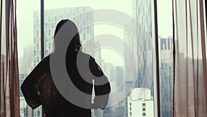 Brunette woman walks to the window stands by the big window looking out apartment on the city buildings. 3840x2160