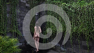 Brunette woman wearing beige bikini taking shower in tropical green Bali garden. Fresh splashes of clean water. Luxury
