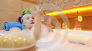 Brunette woman wearing face mask taps on her smartphone in foamy bathtub