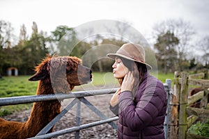 Brunette woman wearing a hat pretending to kiss a brown alpaca behind a fence