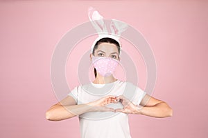 Brunette woman wearing heart print protective face mask and Bunny ears. Girl in white t-shirt on pink background. Easter or