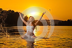 brunette woman in wet white lingerie posing in river water with sunset on background. Young female at the beach in twilight