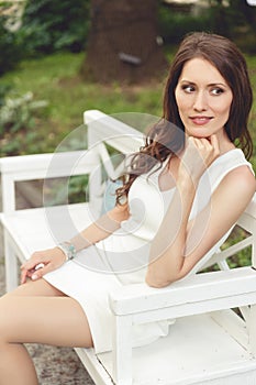 Brunette woman in white dress sitting on bench in park