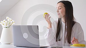 A brunette woman works from home at the computer and eats an apple.