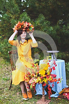 Brunette woman in yellow dress, maple leaves wreath on wooden chair by round table with blue tablecloth. Autumn harvest, nature