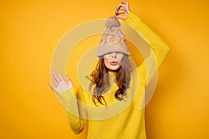 A brunette in a yellow sweater stands on a yellow background with a cap pulled over her face and pulls up for a ponpon