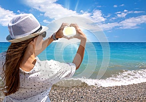 Brunette young girl holding hands in heart shape framing setting sun on sea beach