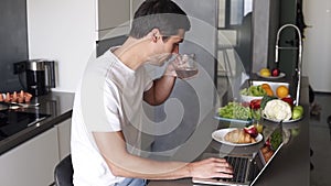 Brunette young man sit and use laptop work in the morning at home in kitchen counter. Man checking e-mails, social media