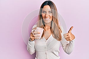 Brunette young woman drinking a cup of coffee smiling happy and positive, thumb up doing excellent and approval sign