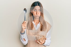 Brunette young woman eating healthy whole grain cereals with spoon puffing cheeks with funny face