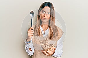 Brunette young woman eating healthy whole grain cereals with spoon relaxed with serious expression on face