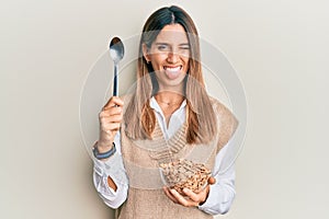 Brunette young woman eating healthy whole grain cereals with spoon sticking tongue out happy with funny expression
