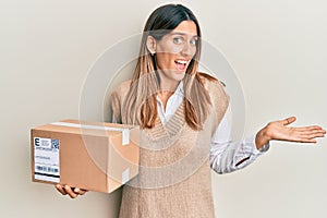 Brunette young woman holding delivery package celebrating achievement with happy smile and winner expression with raised hand