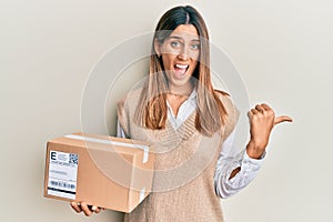 Brunette young woman holding delivery package pointing thumb up to the side smiling happy with open mouth