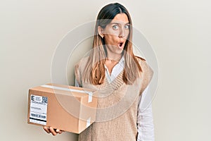 Brunette young woman holding delivery package scared and amazed with open mouth for surprise, disbelief face