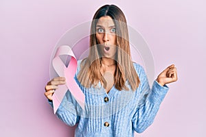 Brunette young woman holding pink cancer ribbon scared and amazed with open mouth for surprise, disbelief face