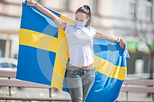 Brunetter standing outdoors looks at the Swedish flag she holds, with a face mask on