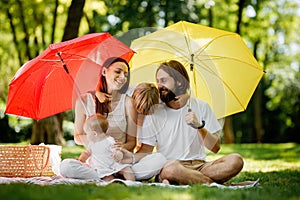 Brunettes parents with two kids have a rest on the lawn under the bright red and yellow umbrellas covering them from the