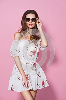 Brunnet girl in floral summer dress and sunglasses posing on pink background. Stylish wavy hairstyle