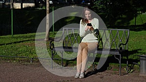 Brunnete woman sitting on bench in park communicating in smartphone