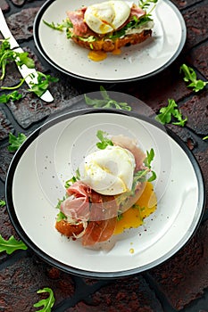 Bruschetta with cream cheese, wilde rucola, parma ham and poached egg served on white plate.