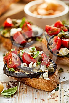 Bruschetta with grilled eggplant, cherry tomatoes, feta cheese, capers and fresh aromatic herbs on a wooden table. Delicious Medit