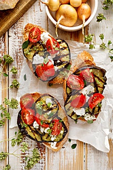 Bruschetta with grilled eggplant, cherry tomatoes, feta cheese, capers and fresh aromatic herbs on a wooden table. Delicious Medit