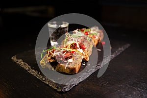 Bruschetta with meat, herbs, and sauce. Restaurant serving on a blackboard on a dark background. A glass of alcohol on