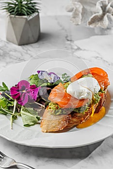 Bruschetta with Poached egg and fresh salmon , vegetarian salad with greens at the white plate on light marble background. Healthy