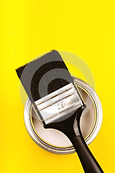 Brush with black handle on open can of paint on yellow background.