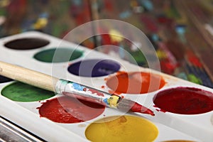 A brush dyed in red paint lies on a set of watercolor paints. Close-up. Sharpness on the tip of the brush.