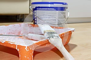 Brush and roller in used orange paint tray, paint can on background. Apartment repair, painting and design concept.