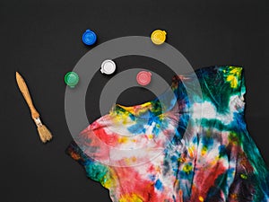 A brush, scattered paint cans, and a tie dye t-shirt on a black background. Flat lay
