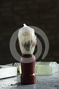 Brush with wooden handle and foam for shave on it, glass bottle of lotion and container for shave cream on the wet grey surface