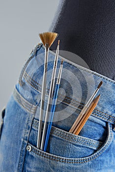 Brushes and pencils in the pocket of the girl`s jeans on a gray background. Artist painting concept