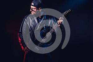 Brutal bearded Heavy metal musician in leather jacket, cap and sunglasses is playing electrical guitar. Shot in a studio
